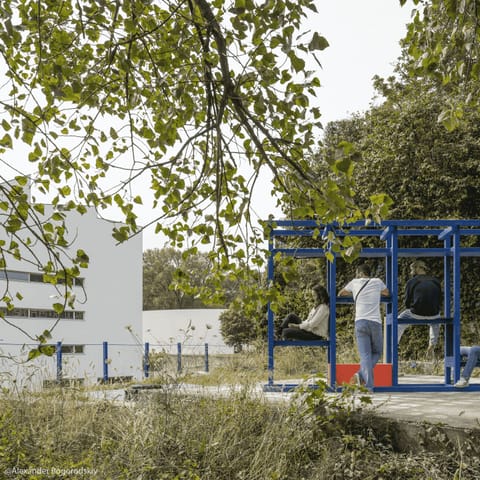 Visitors using the blue steel Petites Folies installation among trees beside a white building in Porto.