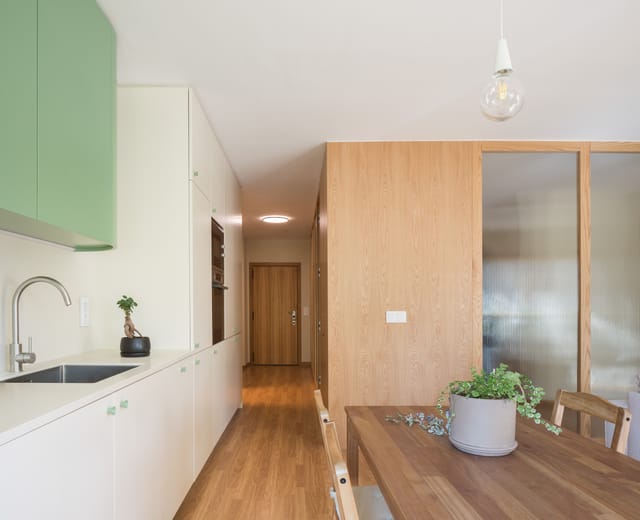 Galley kitchen with white units and green upper cabinets, a dining table and the oak volume beyond.