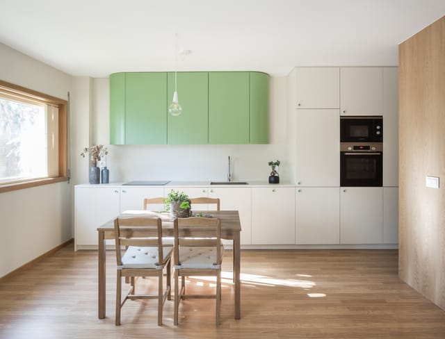 Kitchen and dining area with rounded green wall cabinets, white units and a wooden table by the window.