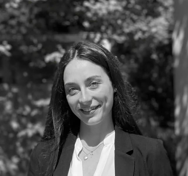 Black-and-white portrait of Marta Maçães Viana smiling, in a dark blazer, in front of foliage.